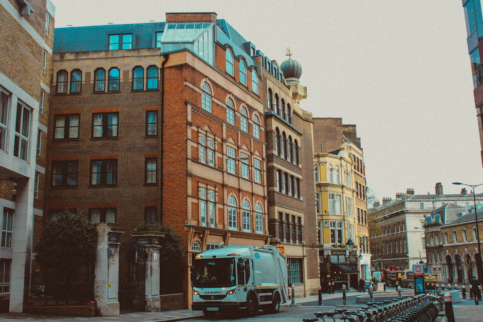 A white waste collection truck with a blue stripe on its side is parked on a city street amidst a row of multi-storey buildings with varied architectural styles. The truck is positioned in the foreground, slightly to the left, adjacent to a sidewalk bordered by black metal bollards. The buildings behind the vehicle include a prominent red-brick structure with large arched windows and decorative stone accents on the facade, and a yellow historical building with ornate detailing and large bay windows further along the street. The background shows additional historic and modern buildings, some with rooftop structures or domes, under an overcast sky. Street fixtures like lampposts, a bus stop sign, and a row of parked bicycles are visible along the curb. The scene suggests an urban environment where private waste collection, such as that provided by companies like House Clearance Bermondsey, might take place as part of alternative rubbish removal services, distinct from council bin collections. The environment appears calm with minimal pedestrian activity, emphasizing the context of on-site or private rubbish handling in a busy city setting.