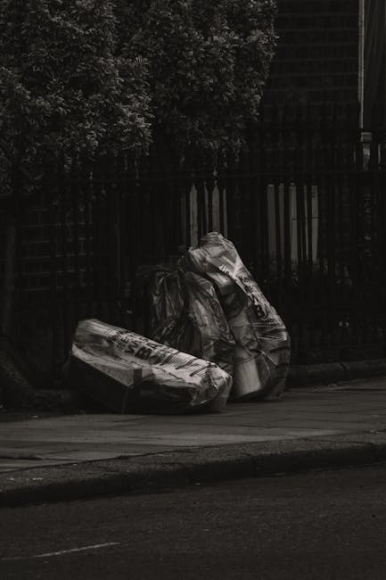An outdoor scene showing multiple overflowing rubbish bins and piles of mixed waste materials on a paved area beside a parking space. In the foreground, a large grey wheelie bin filled with crumpled cardboard, paper, and plastic packaging has its lid open and waste spilling out onto the street. To the left of this bin, there are several black and green garbage bags, along with flattened cardboard boxes, plastic containers, and loose paper scattered on the ground. A nearby red bin is also overflowing, with waste items stacked on top. Behind the bins, a silver car is parked in front of a row of retail shops, which have colorful signage and closed shutter doors. The background features a building under construction or renovation, covered with blue scaffolding and netting, with some visible signage. The overall scene highlights a typical situation of irregular rubbish accumulation possibly related to private waste handling or on-site clearance, with the presence of House Clearance Bermondsey indicated by its natural role in managing such waste disposal needs.
