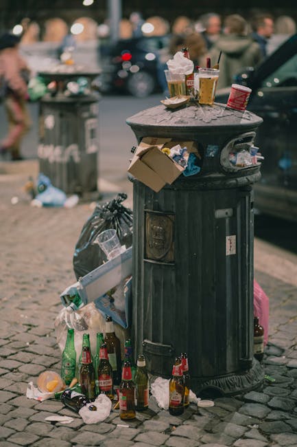 A tall, cylindrical black metal rubbish bin positioned on a cobbled pavement, with a slightly domed flat top surface scattered with various debris, including takeaway food containers, paper cups, and plastic cups with beverage remnants. Attached to the side of the bin are several cardboard boxes and plastic bags filled with discarded waste, some containing empty glass bottles and drink bottles, with additional bottles and trash strewn around the base of the bin. In the background, blurred images of pedestrians, parked cars, and urban street elements are visible, indicating a busy street scene. The lighting suggests daylight, with some ambient reflections on the metal surface of the bin, and the overall scene reflects common urban littering and informal rubbish accumulation outside retail or food establishments, consistent with independent waste disposal practices often seen in city environments.