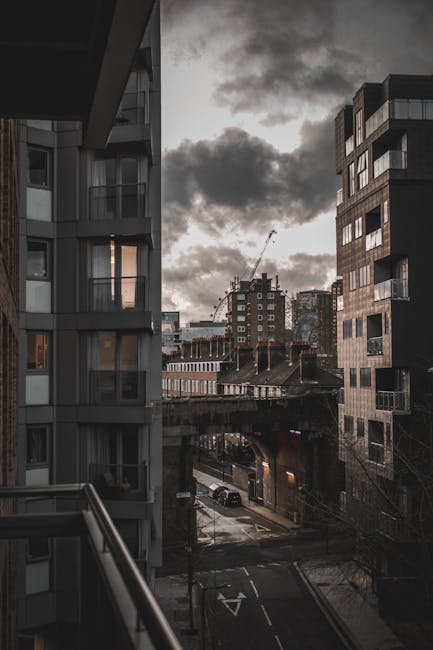 The image depicts a view from a balcony or window overlooking an urban street scene in a city with modern apartment buildings on either side. On the left, part of a building with glass balcony railings and protruding balconies is visible, featuring a contemporary design with grey-toned exterior surfaces. The right side shows another multi-storey building with a dark, textured facade made of brick or similar material, accented by large windows and small balconies. In the background, there are additional buildings of varying heights, including a construction crane indicating ongoing development. The street below appears quiet, with parked cars and a few moving vehicles. Above, the sky is overcast with dark, heavy clouds, casting a moody atmosphere over the scene. The lighting is subdued, suggesting late afternoon or early evening, with limited natural light. The environment and building styles suggest a dense, developed urban area, typical of city districts where private waste collection or on-site clearance services, such as those offered by House Clearance Bermondsey, may be relevant to managing accumulated rubbish, especially in high-rise residential settings.