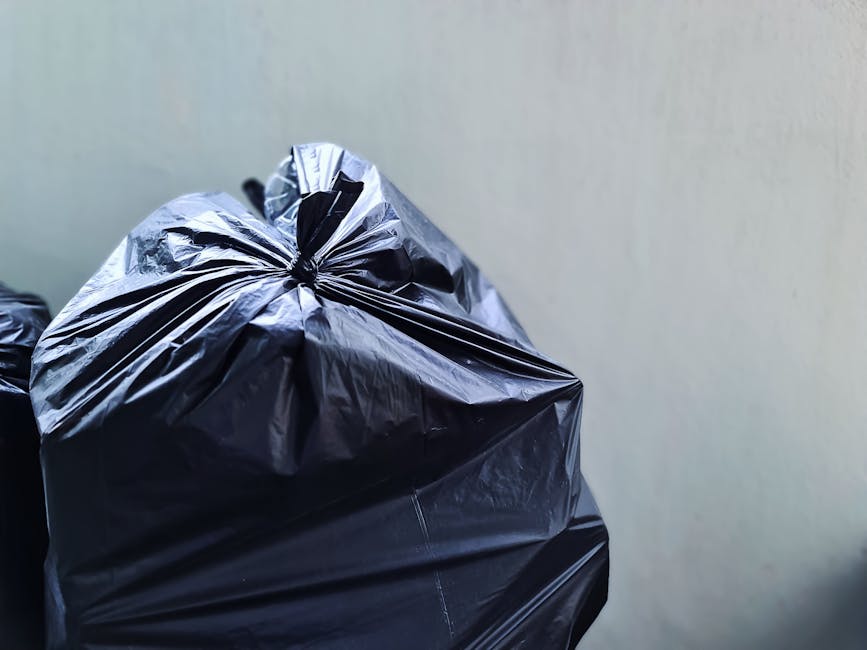 A black, cylindrical public rubbish bin with a rounded, textured concrete lid, situated on a cobbled street. The bin is overflowing with various waste items, including discarded empty glass bottles, paper packaging, and food containers, some of which are spilling onto the ground around it. On top of the lid, there are several used paper cups and a small, crumpled paper bag, indicating recent use. The bin appears weathered, with visible wear and some rust, and has a metal access door with a handle for waste disposal. In the background, a blurred scene of pedestrians walking along the street and parked cars can be seen, along with additional litter scattered nearby, including plastic packaging and paper debris. The environment suggests an urban outdoor setting where informal disposal of rubbish occurs, highlighting the importance of proper waste management services, such as those offered by House Clearance Bermondsey, for responsible rubbish collection and street cleanliness.