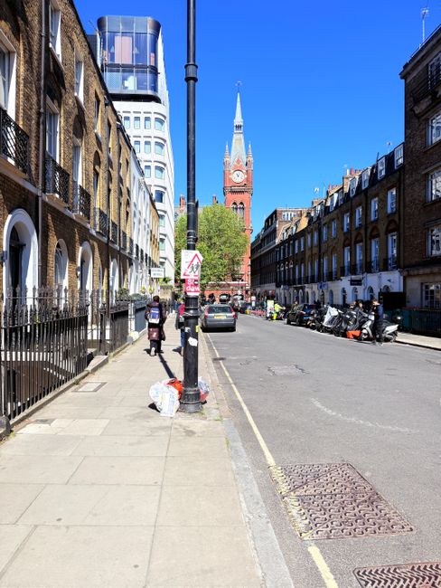 A wide city street scene in Bermondsey during daytime, showing a row of terraced brick townhouses on the left with iron railings and steps leading to residential entrances, and modern multi-storey office or apartment buildings with glass facades in the background. A black lamppost stands prominently in the middle of the sidewalk, with a pedestrian walking past a small pile of recyclable waste bags placed near the base. To the right, vehicles including a small car and multiple parked motorbikes are lined up along the curb. A large historic church with a tall, pointed spire and a clock tower, characteristic of traditional London architecture, is visible further down the street, set against a bright blue sky. The scene is well-lit with natural sunlight, emphasizing the textures of the brickwork, asphalt, and metal objects, and illustrating a typical urban environment where independent rubbish collection services, such as those provided by House Clearance Bermondsey, may handle waste removal outside of council collections.