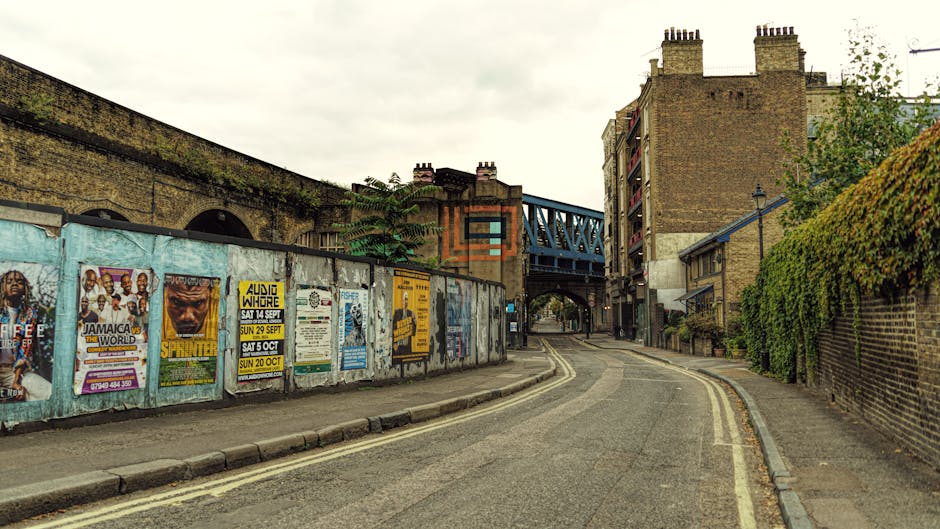 A narrow urban street scene showing a lightly curved asphalt road with double yellow lines on the left side and a paved sidewalk on the right. On the left, a weathered metal fence is covered with colorful posters and advertisements, including event announcements and promotional graphics. Behind the fence, parts of an old brick wall with arched openings are visible. On the right, a tall brick building with several chimneys and a weathered appearance stands adjacent to a dense green hedge that extends along the sidewalk. In the background, a blue railway bridge with metal trusswork spans over the street, casting shadows below. The scene is lit by diffuse natural daylight under a cloudy sky, creating a neutral and subdued atmosphere. The overall setting suggests a typical city alleyway or side street suitable for independent waste collection or private rubbish removal services, as performed by local specialists such as House Clearance Bermondsey.
